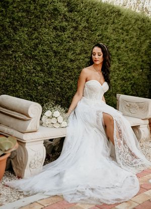 Elegant wedding dress with lace details and thigh-high slit showcased in garden setting with stone bench.