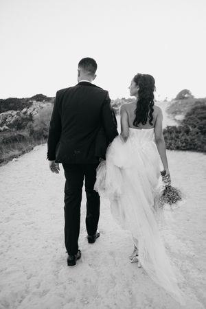 Black and white sequence of newlyweds walking hand in hand along a sandy beach path in their wedding attire.