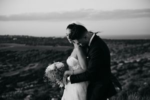 Romantic black and white wedding photo showing couple embracing on hilltop overlooking scenic landscape at sunset.