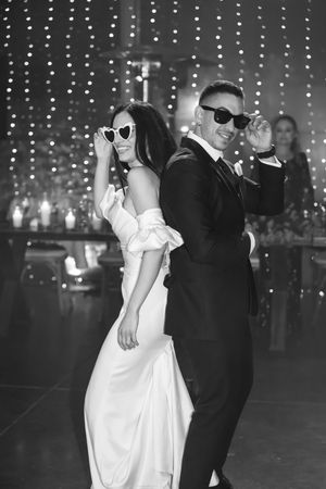 A couple in formal attire dances together at night, with the person in white wearing sunglasses and a flowing dress.
