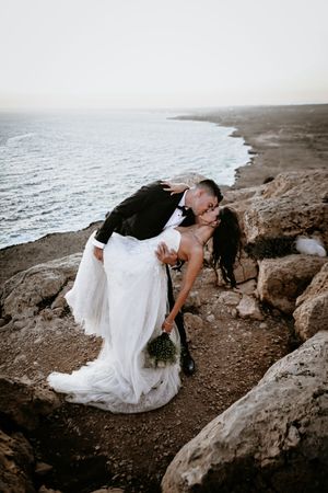 Romantic couple sharing intimate moment during coastal wedding photoshoot on rocky cliffside overlooking misty ocean.