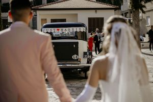 Couple in wedding attire hold hands while looking at vintage black car with guests walking in the distance.