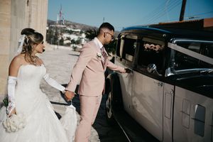 A couple in white and pink formal attire walk together near cars parked outside.