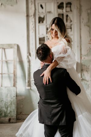 A romantic wedding moment shows an intimate embrace between newlyweds in a rustic indoor setting.