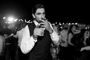 A person in formal attire smoking a cigarette at a nighttime party with blurred lights in the background.