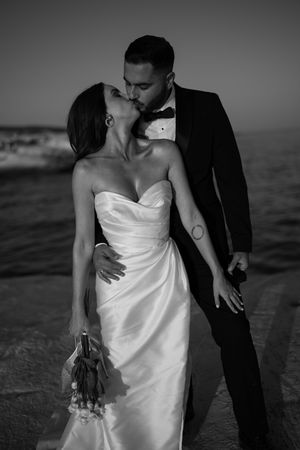 Black and white photo of a bride and groom sharing a kiss by the ocean, bride holding a bouquet in a strapless gown.