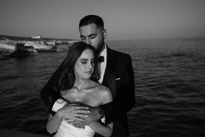 Black and white wedding photo of bride and groom embracing by the sea, groom in suit kissing bride's head.