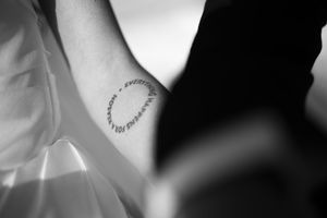 Black and white close-up of a wrist tattoo reading text in a circular design on a wedding day.