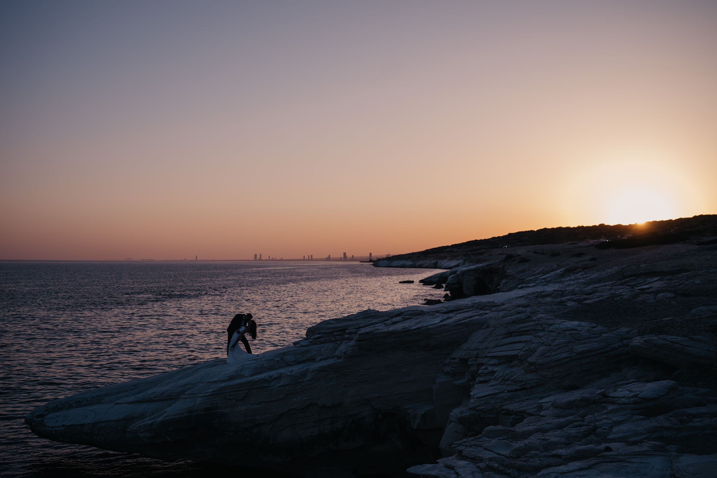 Silhouette of a person on rocky coastal cliffs during a stunning sunset with city skyline in the distance.