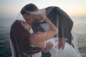 Bride and groom sharing a romantic kiss under a wedding veil by the ocean at sunset.