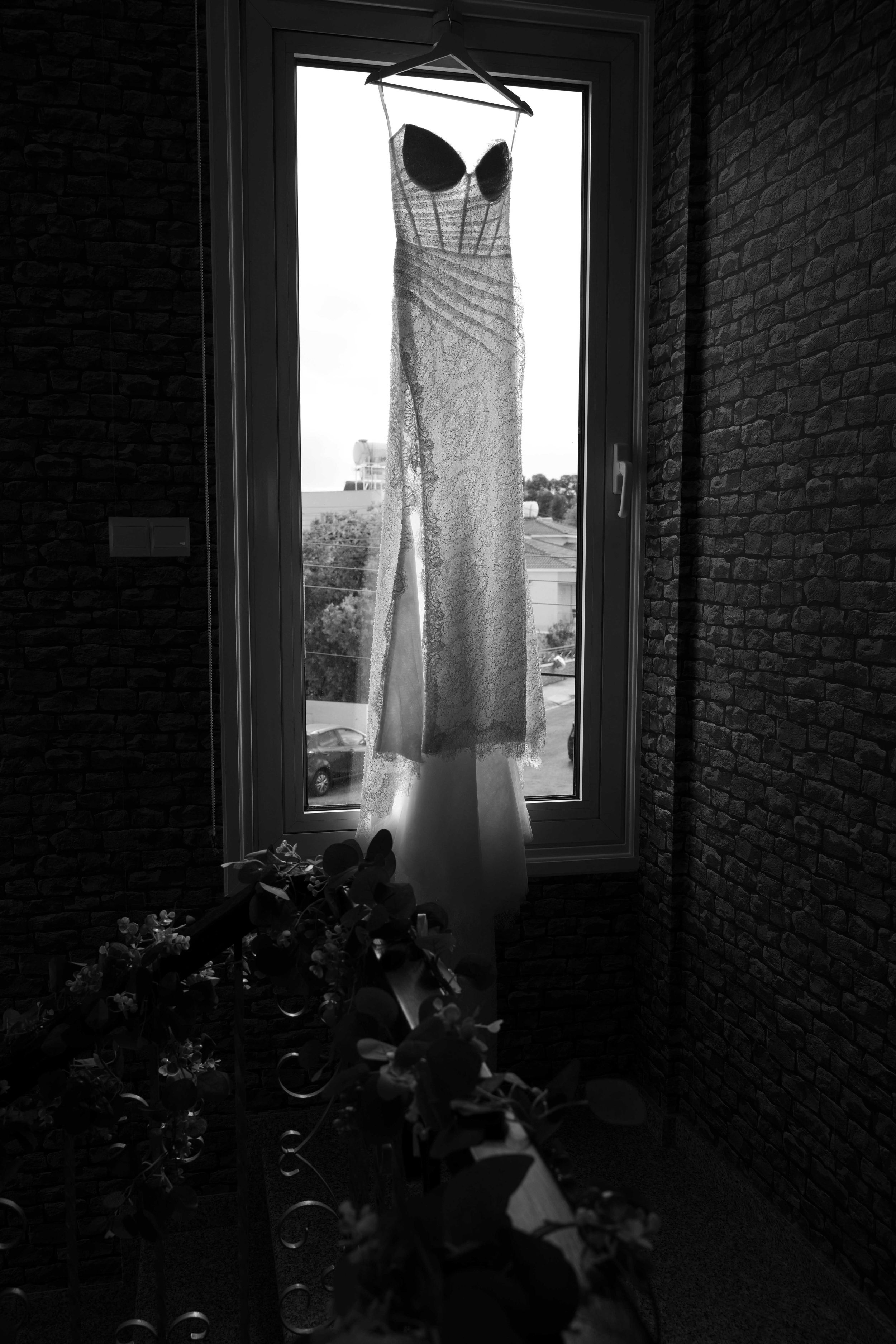 A black and white photograph of a wedding dress hanging in a window frame with backlighting and flowers below.
