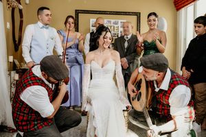 Two people in red plaid kilts kneel to help adjust a white wedding dress while others watch in a hallway.