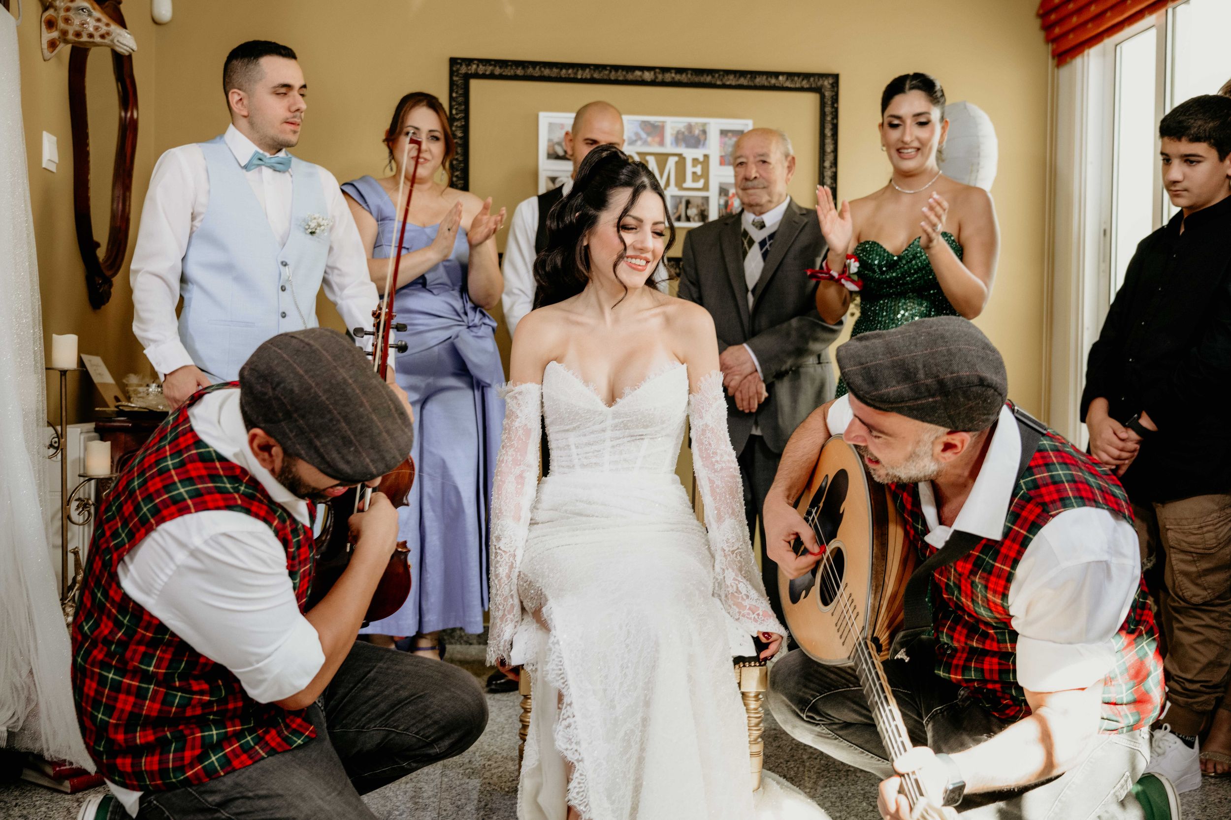 Two people in red plaid kilts kneel to help adjust a white wedding dress while others watch in a hallway.