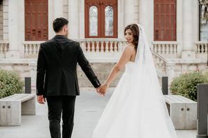 Elegant wedding couple walks along a grand white colonial building with ornate architecture.