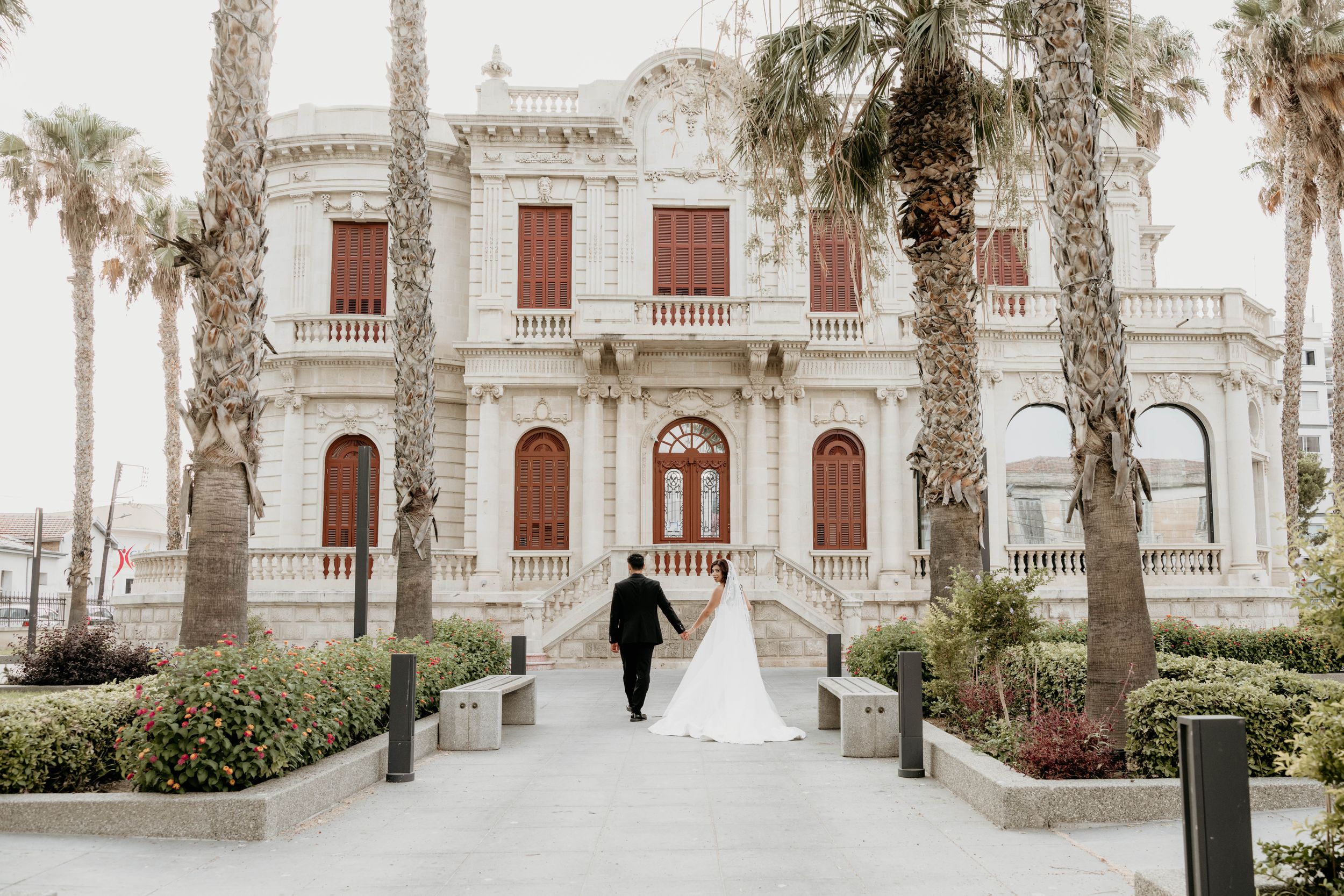 Wedding couple strolls past palm trees and white columns of a historic building.