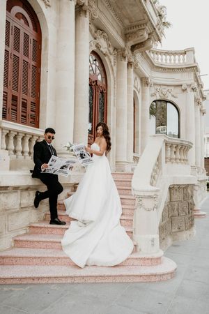 Bride in flowing white dress descends ornate stone steps while groom watches.