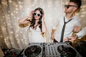Wedding DJ playing music at reception with twinkle lights in background while guests dance and celebrate.