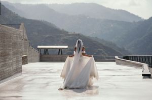A bride in a flowing white dress stands on a rooftop terrace with mountain vistas in the background.