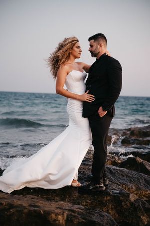 A romantic couple in wedding attire embrace on rocky coastal cliffs as the bride's white dress flows in the ocean breeze.
