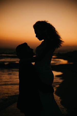 Silhouetted couple embracing on beach at sunset with warm orange sky and ocean waves in background.