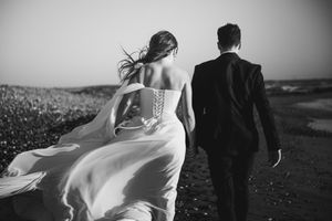 Black and white photo of a couple in wedding attire walking through a scenic outdoor field with flowing dress in wind.