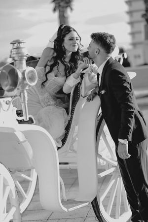 A romantic black and white photo of a couple in wedding attire sharing an intimate moment beside a horse-drawn carriage.