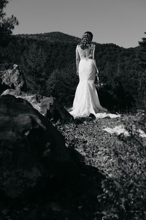 Black and white photo of a bride in a lace back wedding gown standing on a rocky mountain overlook at sunset.