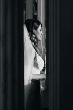 A side profile silhouette of a bride in a white wedding dress and veil against dark curtains in black and white.