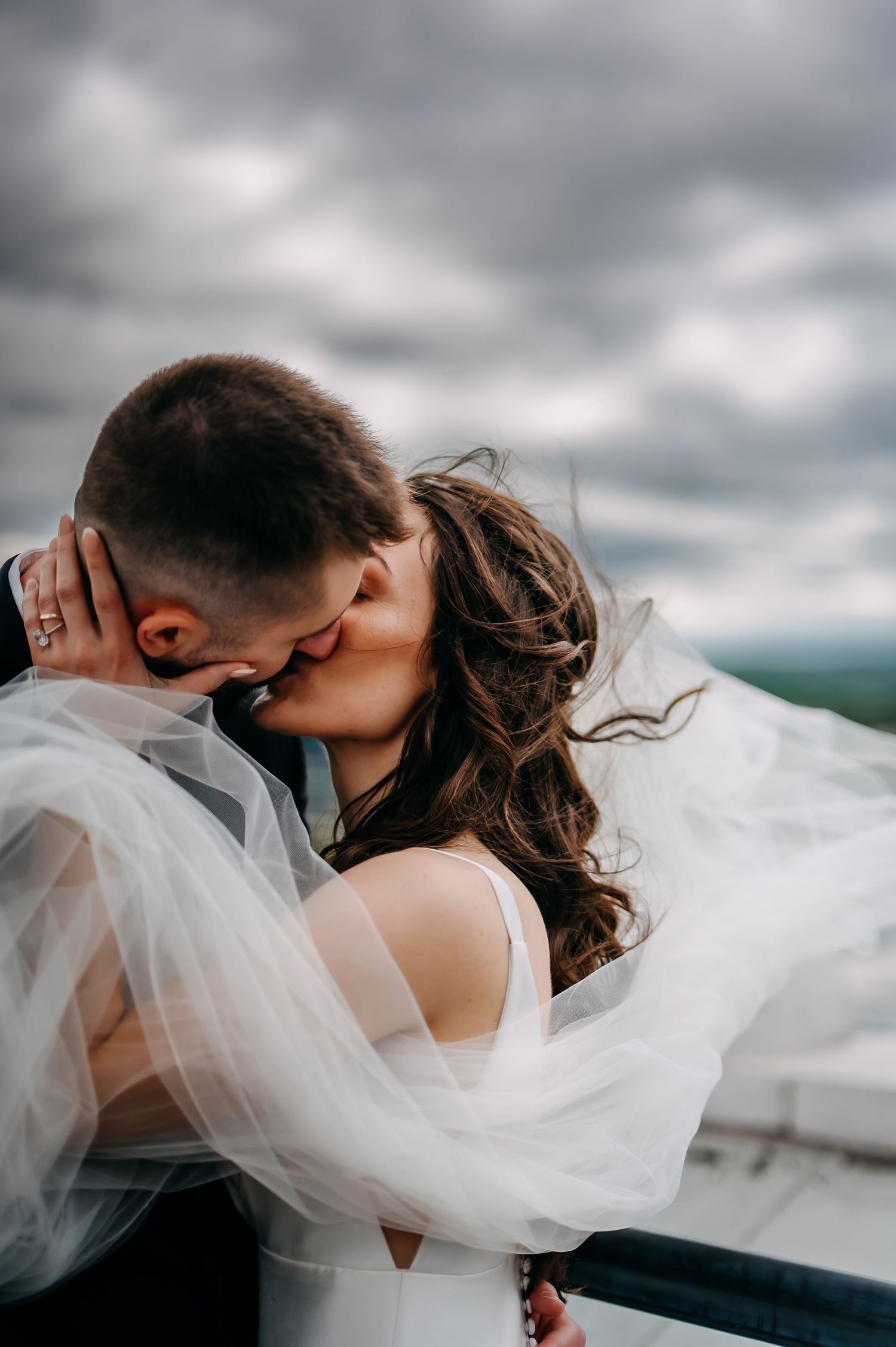 A romantic wedding couple embraces and kisses under dramatic stormy clouds as a delicate white veil flows in the wind.