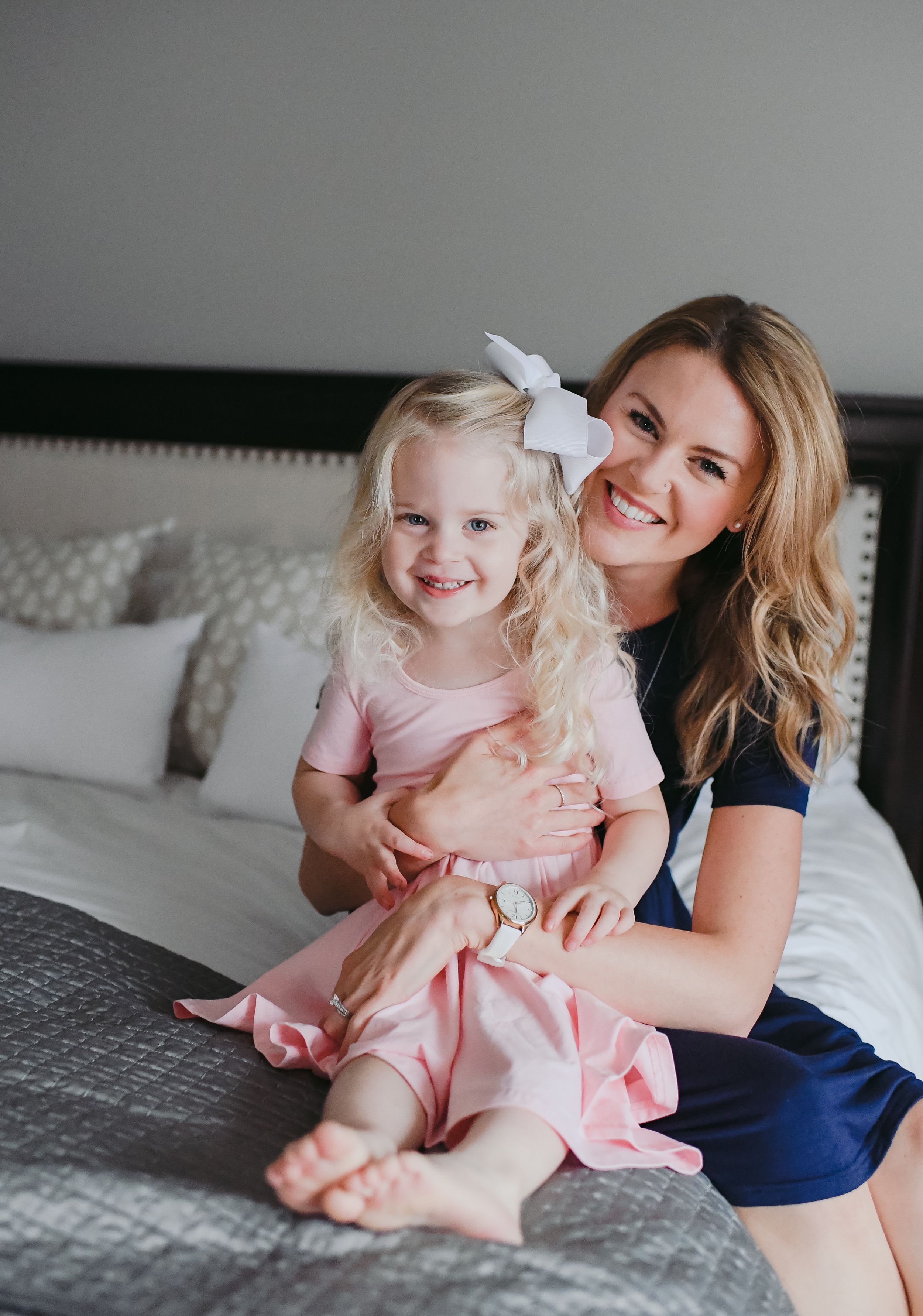 A parent and young child in pink dress sit together smiling on a gray bed.