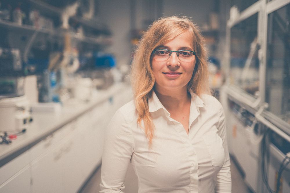 A scientist wearing glasses and a white lab coat stands in a laboratory hallway with equipment visible in background.