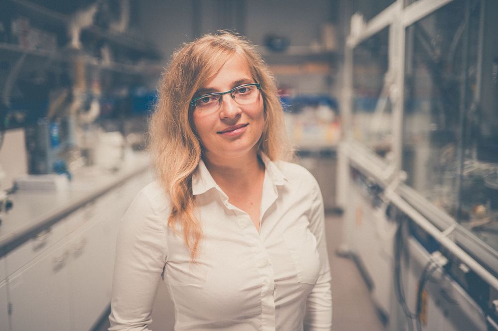 A professional business person smiling while standing against a metal railing backdrop in an office setting.