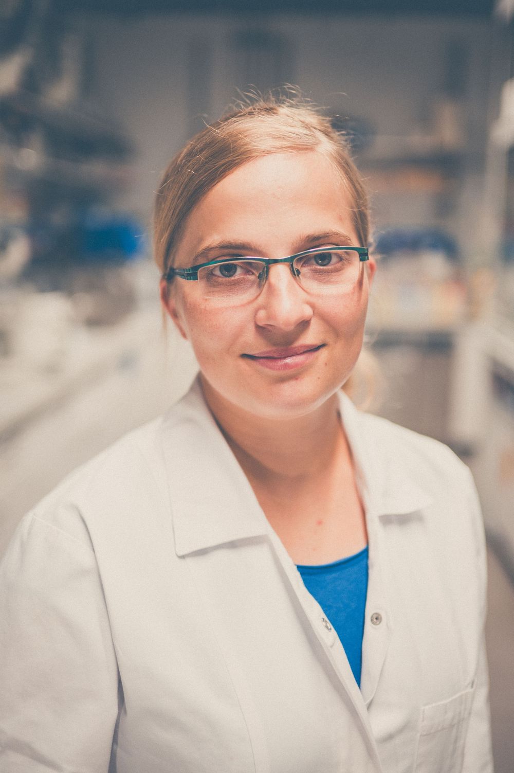 A scientist or medical professional in a white lab coat and glasses smiles warmly in a laboratory setting.