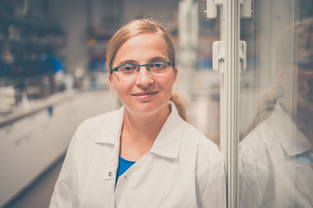 A scientist in a white lab coat and glasses smiles warmly in a laboratory setting.