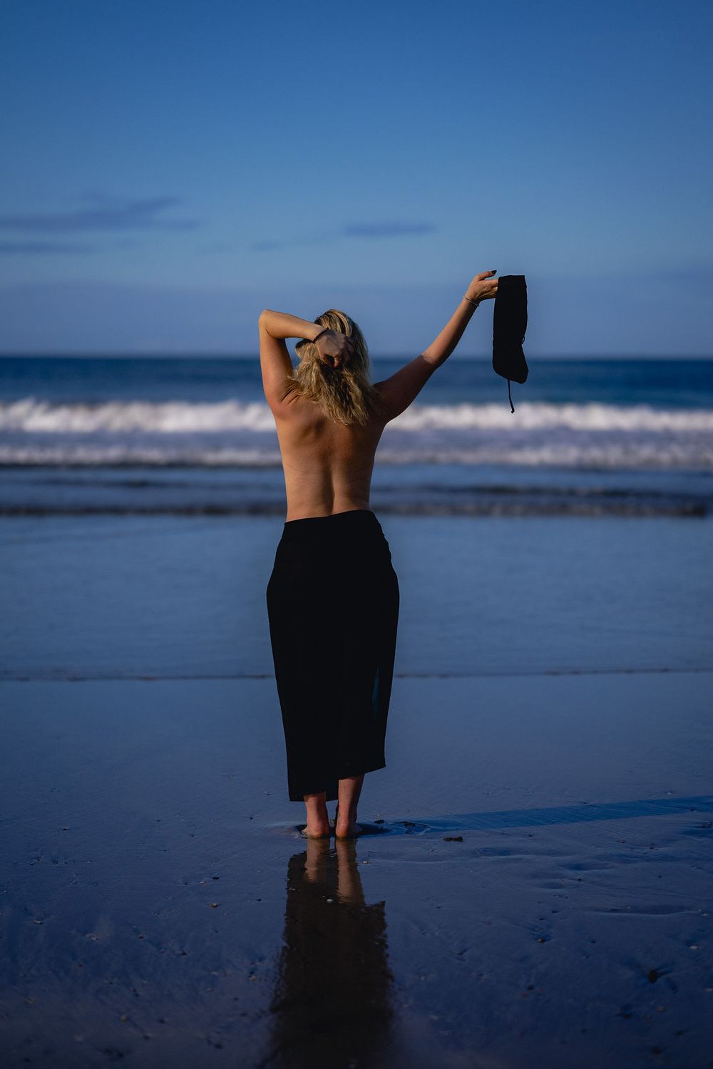 Person undressing on a blue twilight beach shoreline with waves and ocean in the background.