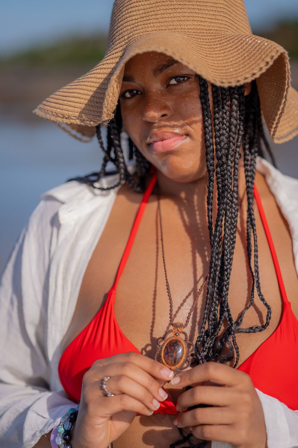 Woman in a straw sun hat and red swimsuit playing with braided hair against a scenic beach backdrop.