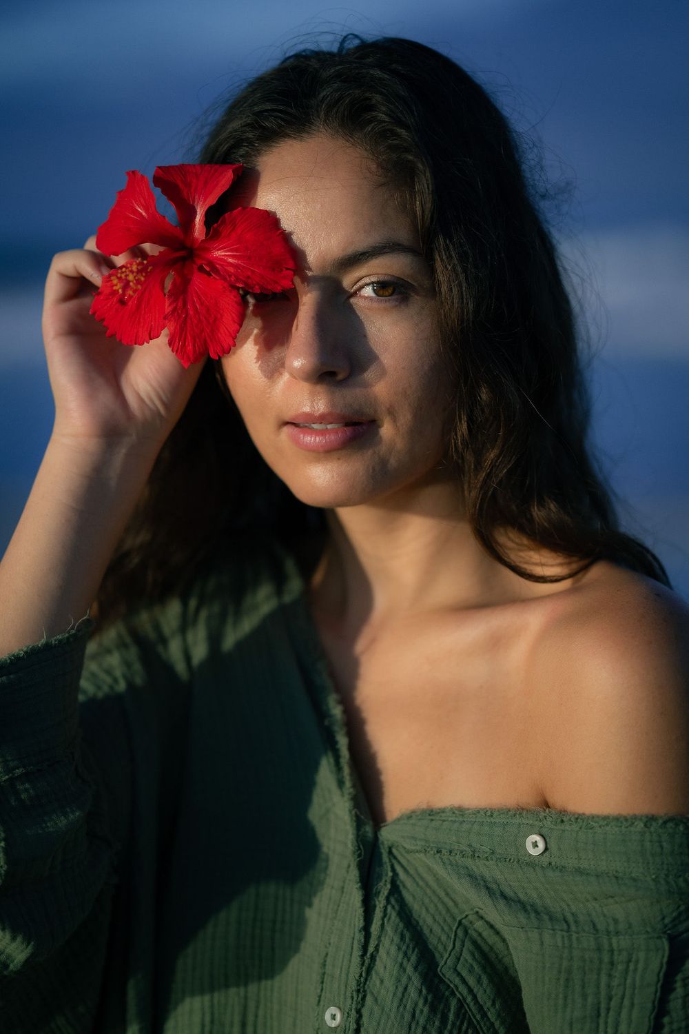 A person in a green off-shoulder top poses with a vibrant red hibiscus flower against a deep blue sky background.