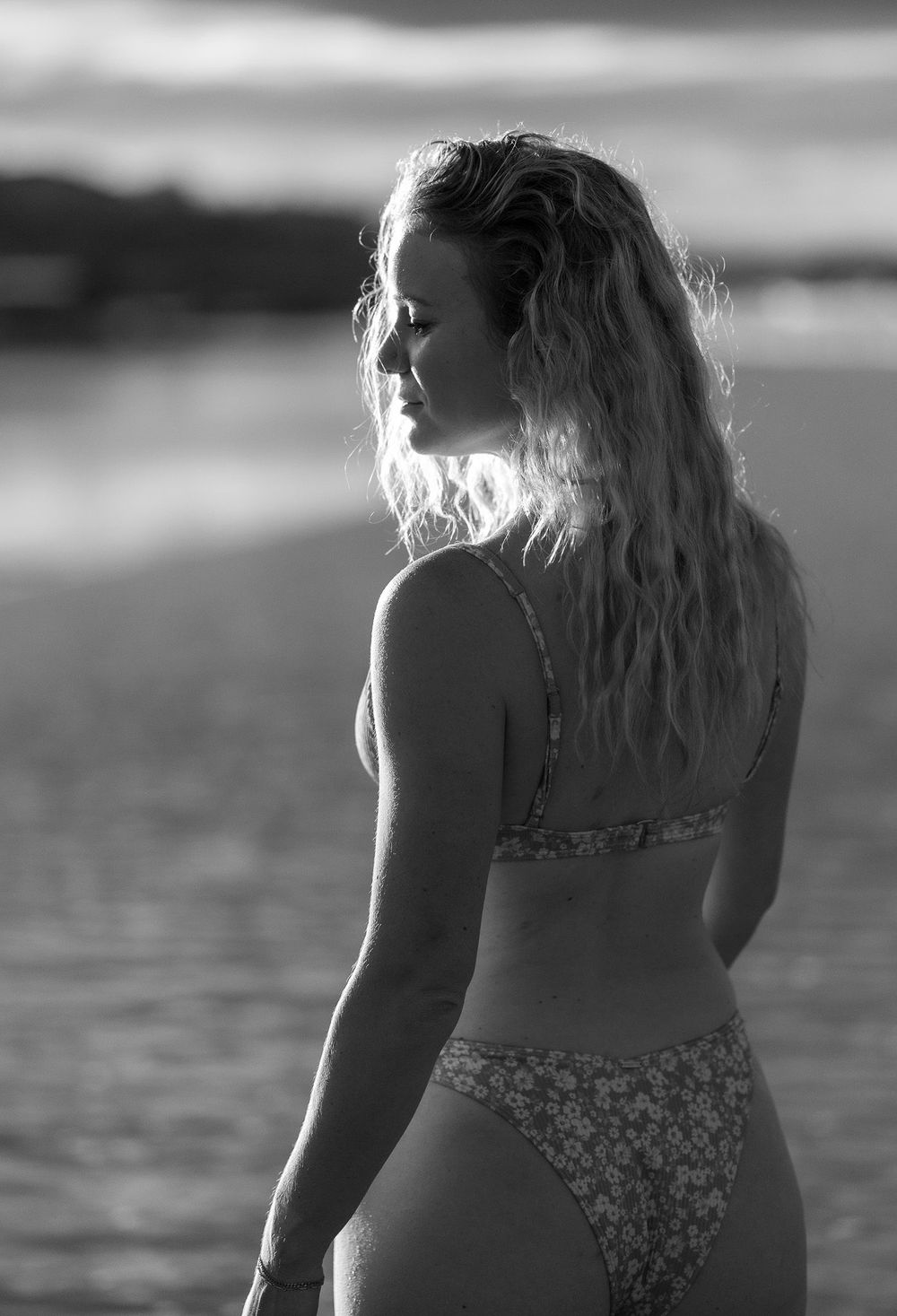 Black and white beach portrait shows a person in floral swimwear looking towards water at sunset.