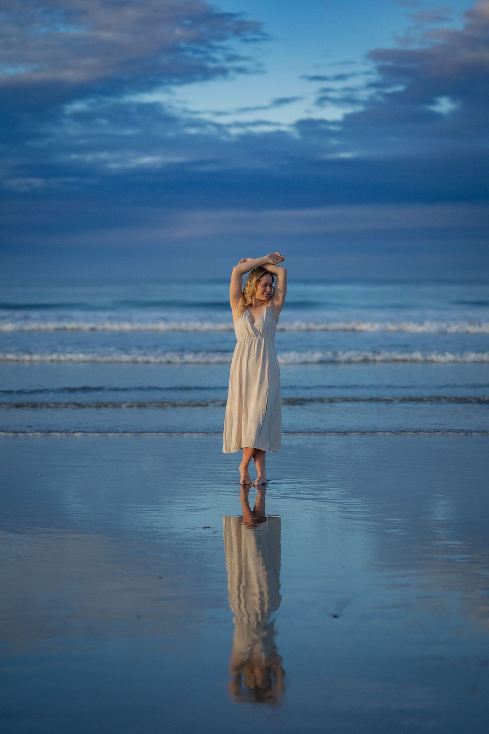A person in a white dress stands reflectively on a wet beach at dusk with dramatic blue cloudy skies above.