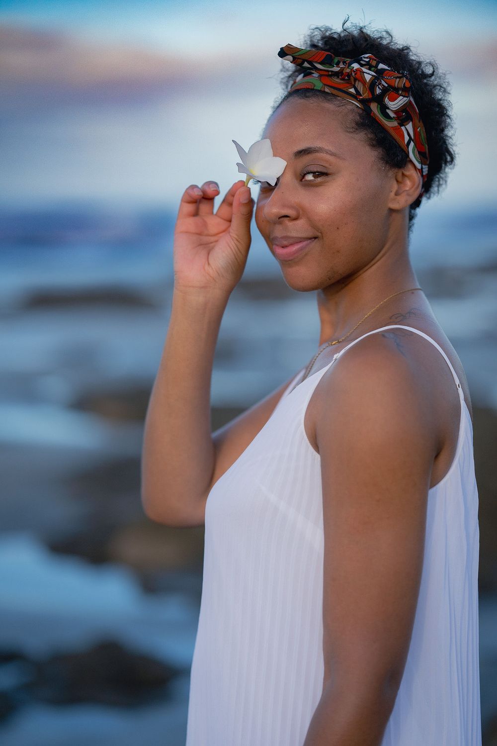 A woman in a white grecian style dress poses against a blue background at sunset.