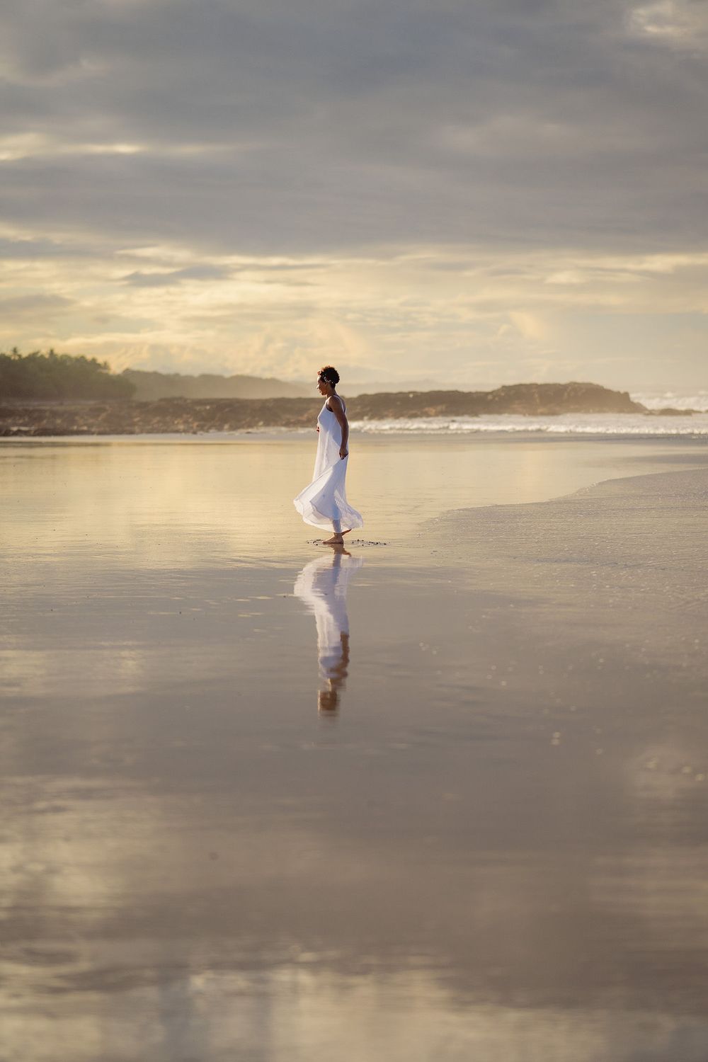 A person in white clothing walks along a misty beach at sunset with dramatic cloudy skies and ocean waves.