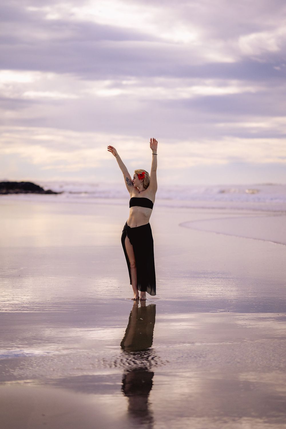 A silhouette dances with raised arms on a reflective beach at sunset with purple cloudy skies overhead.