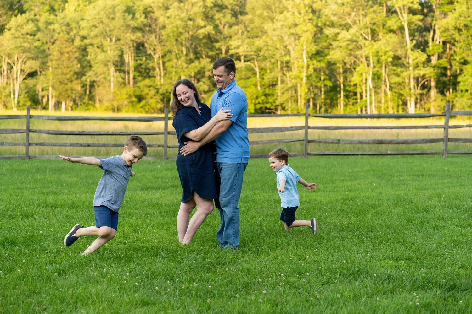 A family enjoying a playful moment together in a lush green field with a wooden fence and trees in the background.