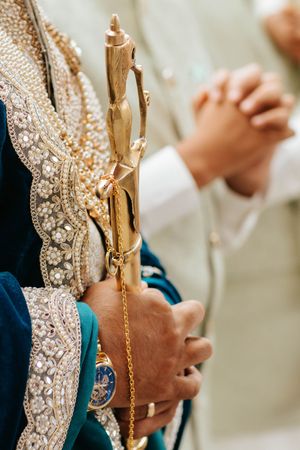 A person wearing traditional ornate Indian attire holds a decorative golden staff with embellished pearls and crystals.