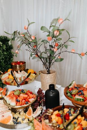 Elegant fruit and cheese display featuring colorful fresh fruits, berries and decorative branches with pink blooms in a vase.