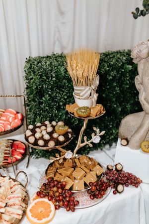 Elegant food display with wheat stalks, grapes, pastries and appetizers on a white tablecloth with greenery backdrop.