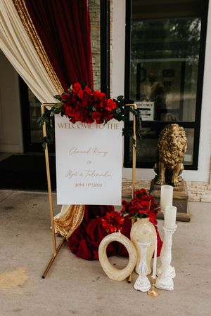 Wedding welcome sign display with red florals, cream drapes, and golden accents for an elegant ceremony entrance.