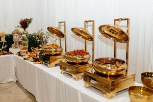 A catering buffet table set with gold warming stations and food displays against a white backdrop.