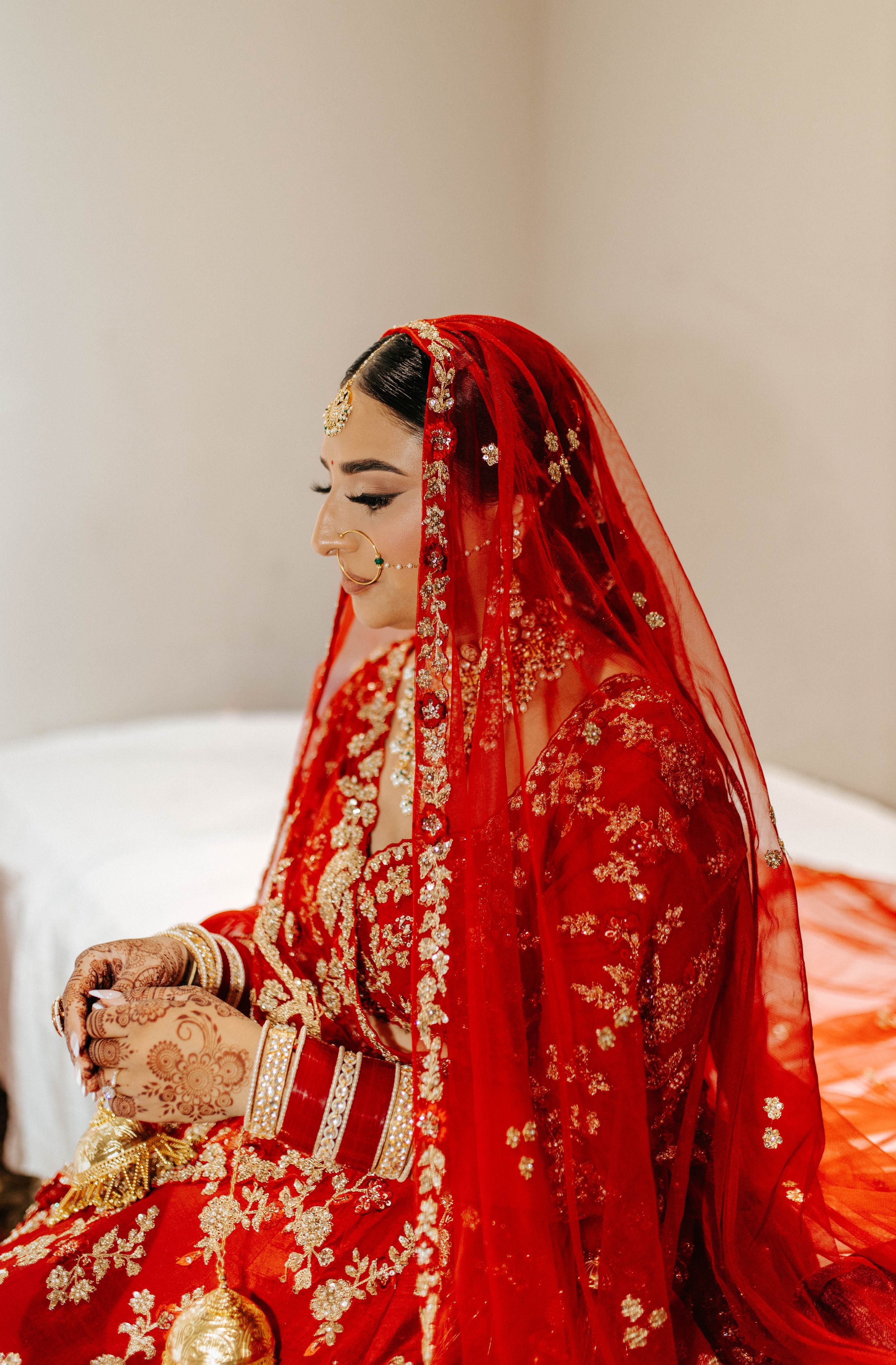 A bride in a traditional red Indian wedding dress with intricate gold embroidery and henna-decorated hands sits in contemplation.