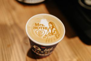 A latte with intricate art design showing figures and text in foam against a wooden table.